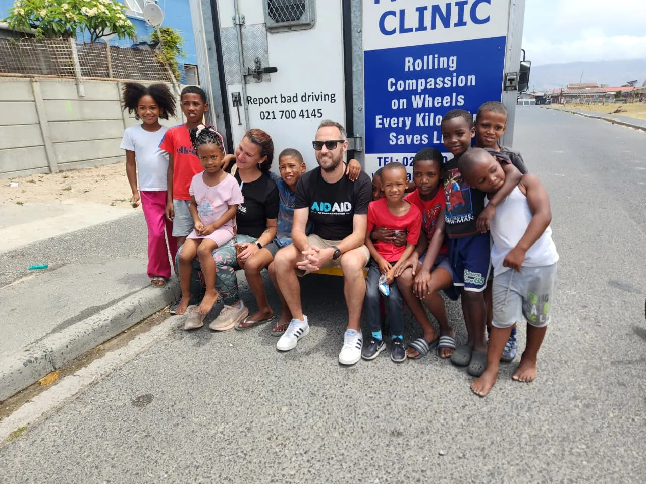 An Aid4Aid volunteer sitting on the tailgate of the mobile clinic truck, surrounded by a crowd of smiling children from the neighbourhood.