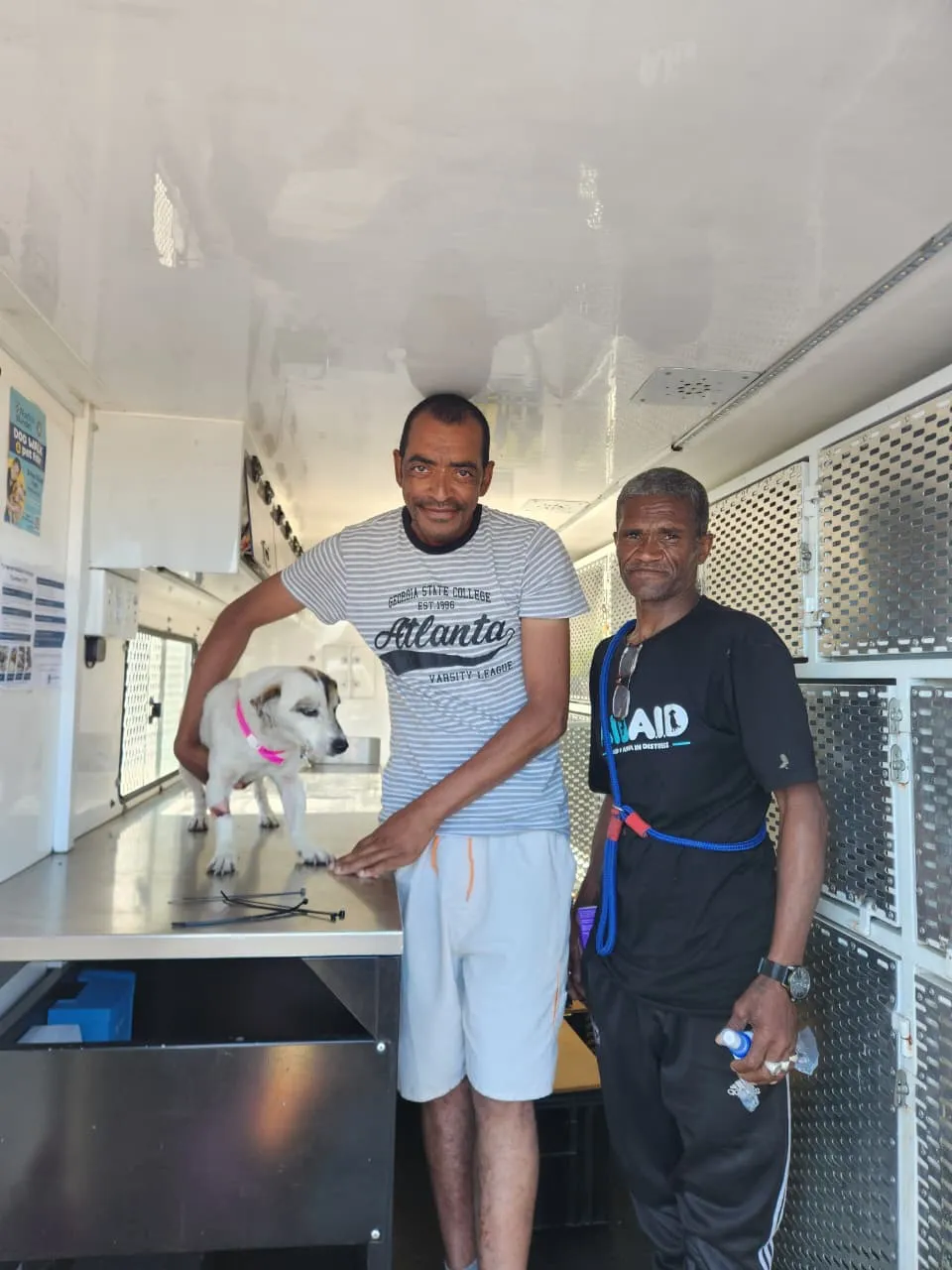 Two men with a small white dog wearing a pink collar on the table inside the mobile clinic, one in an Aid4Aid shirt with a slip lead over his shoulders.
