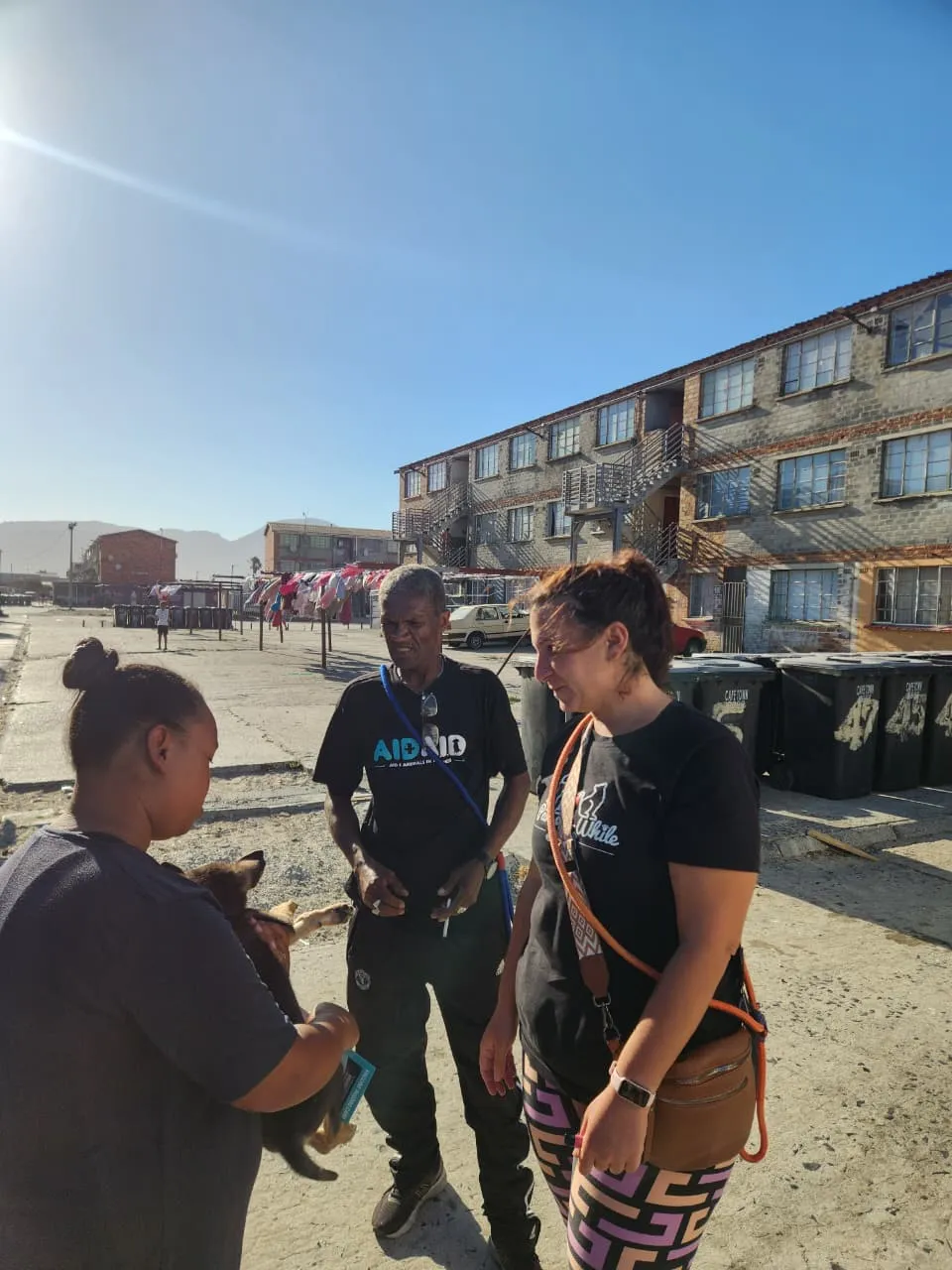 An Aid4Aid field worker and a colleague in a Paws-a-While shirt talking with a resident holding a puppy, on a street between blocks of flats.